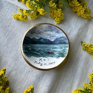 May include: A small, round, hand-painted ceramic dish with a landscape scene of a lake and mountains. The dish has a gold rim and the text "K ❤️ T July 13, 2024". Yellow flowers are arranged around the dish.