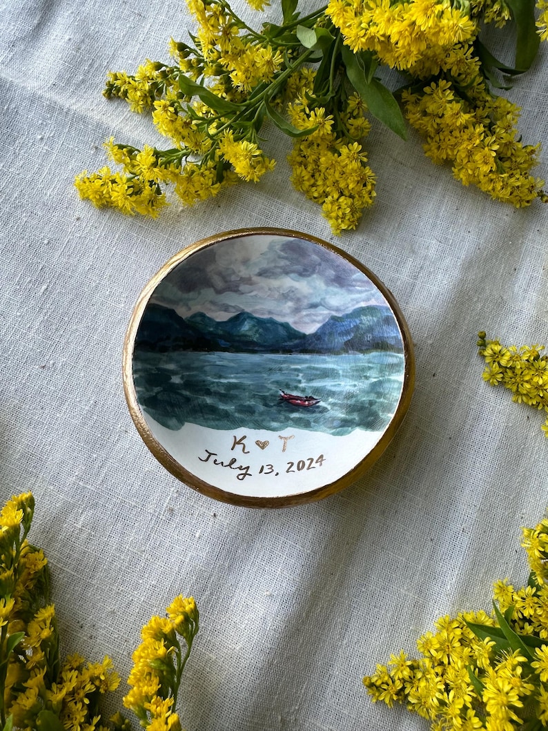 May include: A small, round, hand-painted ceramic dish with a landscape scene of a lake and mountains. The dish has a gold rim and the text "K ❤️ T July 13, 2024". Yellow flowers are arranged around the dish.