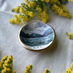 May include: A small, round, wooden trinket dish with a hand-painted seascape scene. The interior features a painting of a boat on a wavy sea with mountains in the background. The dish has a gold rim and the initials "K ❤️ T" are painted on the bottom.