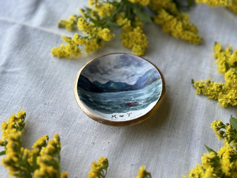 May include: A small, round, wooden trinket dish with a hand-painted seascape scene. The interior features a painting of a boat on a wavy sea with mountains in the background. The dish has a gold rim and the initials "K ❤️ T" are painted on the bottom.