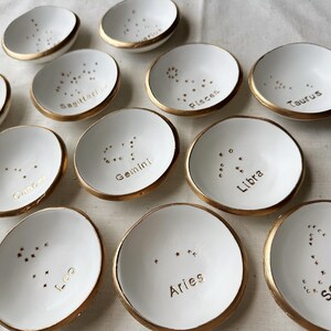 Puede incluir: Doce platos de cer&aacute;mica blanca con bordes dorados y constelaciones pintadas en oro. Cada plato tiene un signo del zod&iacute;aco escrito en letras doradas. Los signos son: Sagitario, Piscis, Tauro, C&aacute;ncer, G&eacute;minis, Libra, Leo, Aries, Escorpio, Capricornio, Acuario y Virgo.