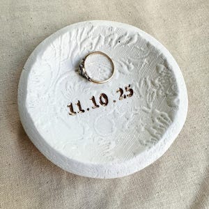 Puede incluir: Un plato redondo de cer&aacute;mica blanca para anillos con un patr&oacute;n floral texturizado y la fecha "11.10.25" en letras doradas. Un anillo de oro con una peque&ntilde;a gema se encuentra en el centro.