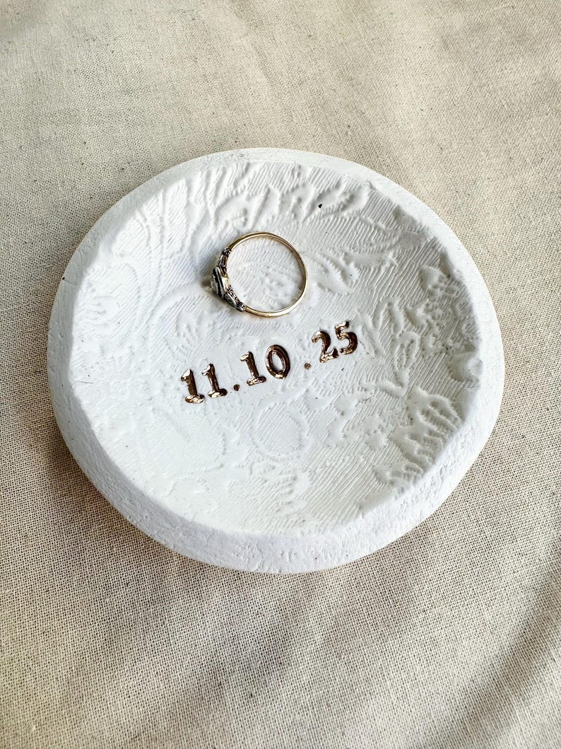 Puede incluir: Un plato redondo de cer&aacute;mica blanca para anillos con un patr&oacute;n floral texturizado y la fecha "11.10.25" en letras doradas. Un anillo de oro con una peque&ntilde;a gema se encuentra en el centro.