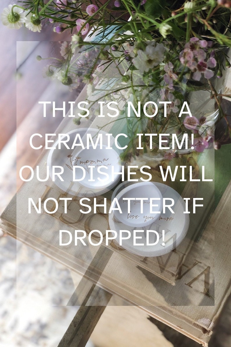 May include: Two white ceramic dishes with handwritten messages. The top dish says "momma dishes" and the bottom dish says "love you more". The dishes are sitting on a stack of books with the text "THIS IS NOT A CERAMIC ITEM! OUR DISHES WILL NOT SHATTER IF DROPPED!" printed on the top book.