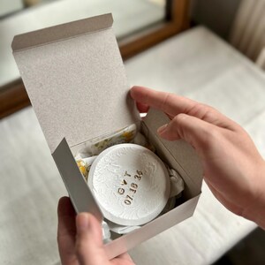 Puede incluir: Un objeto decorativo blanco y redondo con motivos florales y texto dorado dentro de una caja de cart&oacute;n gris. El texto dice "GVT 07.18.26". La caja est&aacute; abierta, revelando el art&iacute;culo y su embalaje.