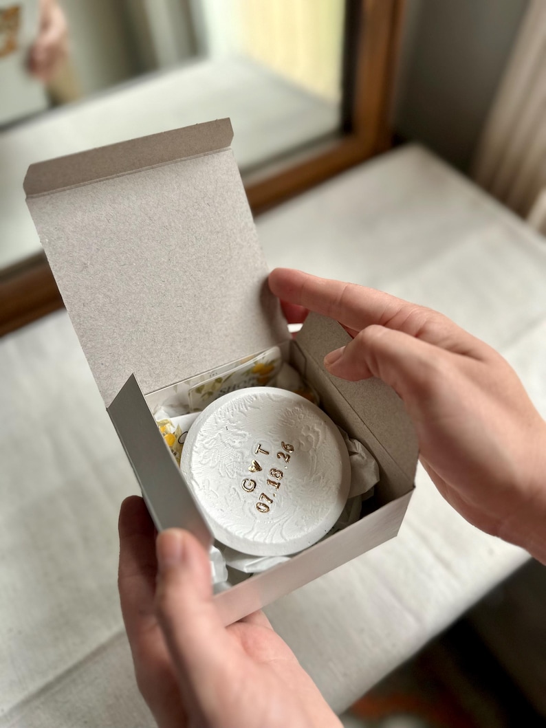 Puede incluir: Un objeto decorativo blanco y redondo con motivos florales y texto dorado dentro de una caja de cart&oacute;n gris. El texto dice "GVT 07.18.26". La caja est&aacute; abierta, revelando el art&iacute;culo y su embalaje.