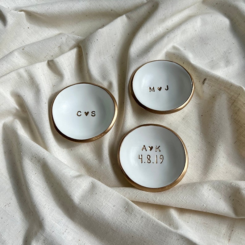 May include: Three white ceramic ring dishes with gold rims. Each dish has a different inscription: "C&hearts;S", "M&hearts;J", and "A&hearts;K 4.8.19".