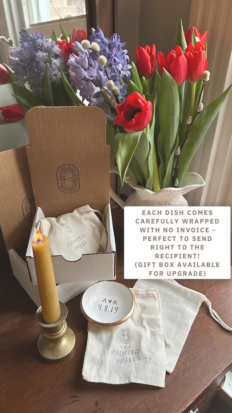Puede incluir: Un peque&ntilde;o plato de cer&aacute;mica blanco con borde dorado, personalizado con iniciales y una fecha, se encuentra cerca de una vela y cajas de regalo. Las cajas contienen peque&ntilde;as bolsas de tela. Un ramo de tulipanes rojos e jacintos morados.