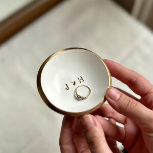 May include: A small, white ceramic ring dish with a gold rim and gold lettering. The dish holds a gold ring with a decorative silver design. The initials "J & H" are stamped in gold on the dish.