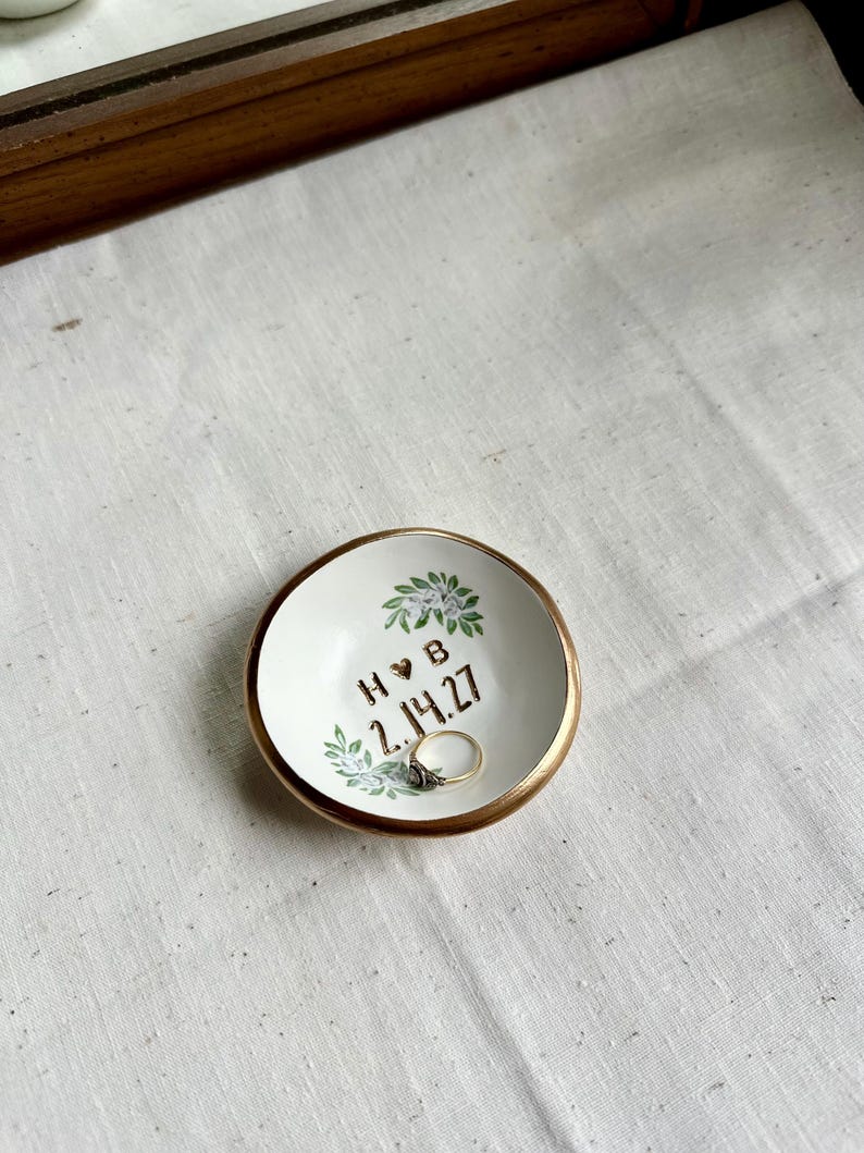 May include: A small, white ceramic ring dish with a gold rim. The dish is decorated with floral accents and the initials "H & B" with a heart symbol, and the date "2.14.27". A ring rests inside the dish.