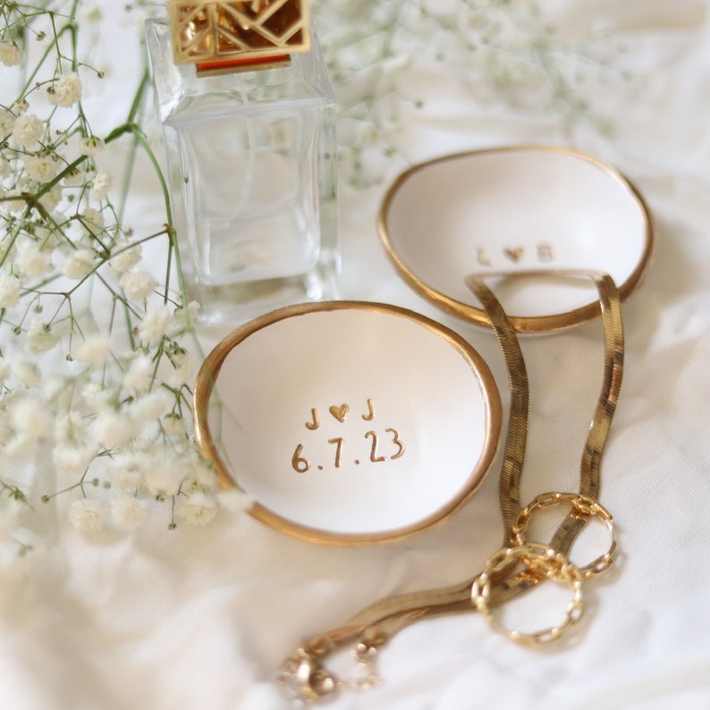 May include: Two white ceramic ring dishes with gold rims. The dish on the left has the inscription "J &hearts; J 6.7.23" and the dish on the right has the inscription "LOVE". A gold chain necklace is lying on the table next to the dishes.