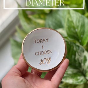 Puede incluir: Un peque&ntilde;o plato de cer&aacute;mica blanco con borde dorado, de aproximadamente 7,6 cm de di&aacute;metro. El plato presenta las palabras "TODAY I CHOOSE JOY" en letras doradas. El fondo es una vista borrosa de follaje verde.