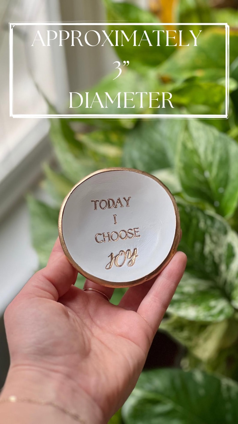 Puede incluir: Un peque&ntilde;o plato de cer&aacute;mica blanco con borde dorado, de aproximadamente 7,6 cm de di&aacute;metro. El plato presenta las palabras "TODAY I CHOOSE JOY" en letras doradas. El fondo es una vista borrosa de follaje verde.