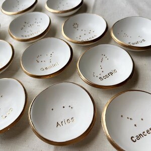 Puede incluir: Conjunto de 12 platos de anillo de cer&aacute;mica blanca con bordes dorados. Cada plato presenta una constelaci&oacute;n zodiacal diferente con su nombre correspondiente en letras doradas. Las constelaciones incluyen Aries, Tauro, G&eacute;minis, C&aacute;ncer, Leo, Virgo, Libra, Escorpio, Sagitario, Capricornio, Acuario y Piscis.