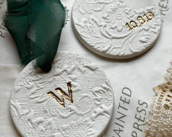 May include: Two white, round ornaments with embossed floral designs. One has the gold letter "W", the other has the gold numbers "10.3.19". Each has a red or green ribbon for hanging. The background includes the text "PAINTED PRESS".