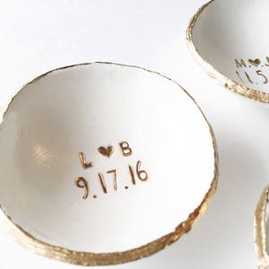 Puede incluir: Dos platos de anillos de cer&aacute;mica blanca con borde dorado. El plato de la izquierda tiene las iniciales "L&hearts;B" y la fecha "9.17.16" inscritas en oro. El plato de la derecha tiene las iniciales "M&hearts;J" y la fecha "11.5.16" inscritas en oro.