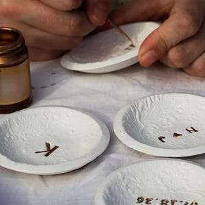 Puede incluir: Platos de cer&aacute;mica blanca texturizada con letras y dise&ntilde;os dorados. Un plato tiene la letra "K", otro "J & N" y un tercero una fecha e iniciales. Una persona est&aacute; pintando un plato con un pincel peque&ntilde;o.
