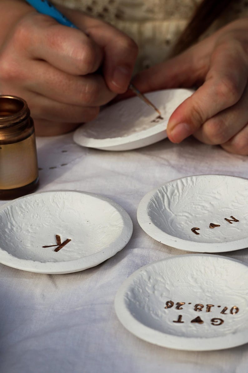 Puede incluir: Platos de cer&aacute;mica blanca texturizada con letras y dise&ntilde;os dorados. Un plato tiene la letra "K", otro "J & N" y un tercero una fecha e iniciales. Una persona est&aacute; pintando un plato con un pincel peque&ntilde;o.