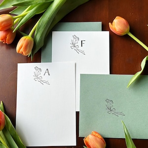 May include: Three white note cards with a floral design and a letter on each card. The letters are A, F, and a blank card. The cards are on a brown wooden surface with orange tulips.