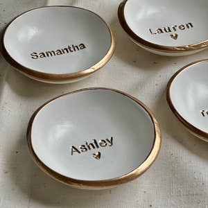 May include: Four white ceramic ring dishes with gold trim. Each dish has a name written on it: Samantha, Lauren, Ashley, and Reese.