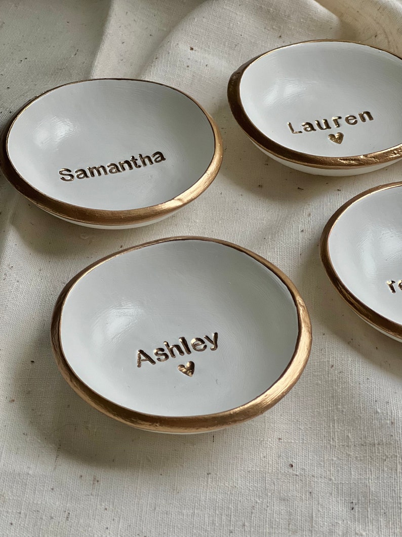 May include: Four white ceramic ring dishes with gold trim. Each dish has a name written on it: Samantha, Lauren, Ashley, and Reese.