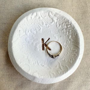 Puede incluir: Un plato de cer&aacute;mica blanco, redondo y repujado con las letras "KO" y un anillo dorado. El plato tiene un dise&ntilde;o floral y est&aacute; sobre una superficie de lino neutro. El anillo es dorado con una banda oscura estampada.