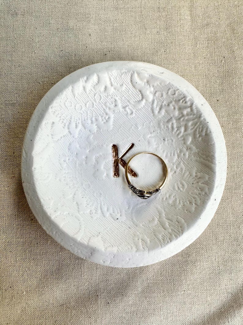 Puede incluir: Un plato de cer&aacute;mica blanco, redondo y repujado con las letras "KO" y un anillo dorado. El plato tiene un dise&ntilde;o floral y est&aacute; sobre una superficie de lino neutro. El anillo es dorado con una banda oscura estampada.