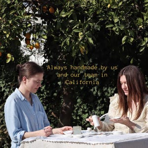 Puede incluir: Dos personas sentadas a una mesa puesta para una comida al aire libre, bajo un &aacute;rbol con naranjas. La mesa est&aacute; cubierta con un mantel blanco. El texto "Always handmade by us and our team in California" es visible.