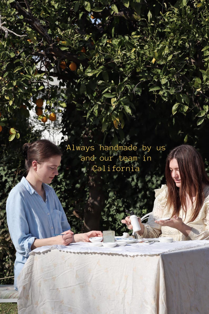 Puede incluir: Dos personas sentadas a una mesa puesta para una comida al aire libre, bajo un &aacute;rbol con naranjas. La mesa est&aacute; cubierta con un mantel blanco. El texto "Always handmade by us and our team in California" es visible.