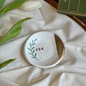 May include: White ceramic ring dish with a gold rim and a green leaf design. The dish has the initials "J & K" written in gold on the front.