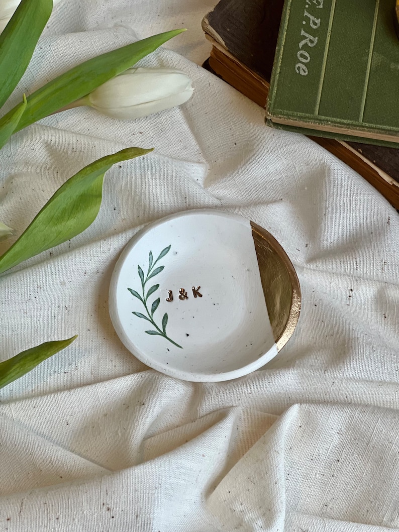 May include: White ceramic ring dish with a gold rim and a green leaf design. The dish has the initials "J & K" written in gold on the front.