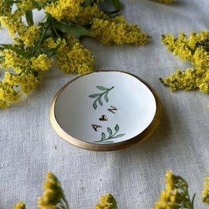 Puede incluir: Plato de cer&aacute;mica blanca para anillos con borde dorado. El plato est&aacute; decorado con hojas verdes y las letras A y N con un coraz&oacute;n entre ellas.