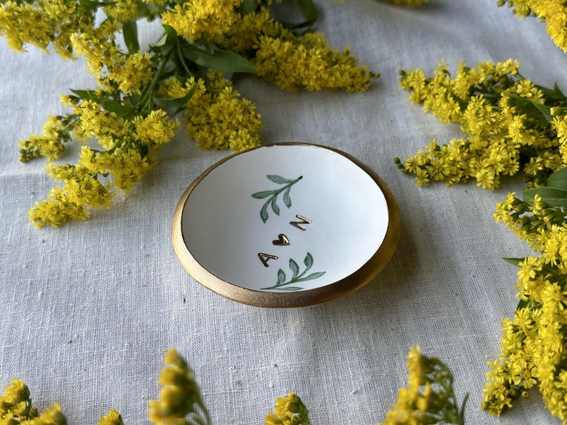 Puede incluir: Plato de cer&aacute;mica blanca para anillos con borde dorado. El plato est&aacute; decorado con hojas verdes y las letras A y N con un coraz&oacute;n entre ellas.