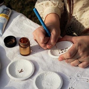 Puede incluir: Una persona est&aacute; rotulando a mano un peque&ntilde;o plato de cer&aacute;mica blanco con un bol&iacute;grafo azul. Otros platos blancos con dise&ntilde;os en relieve y un peque&ntilde;o tarro dorado est&aacute;n sobre un mantel blanco. Tambi&eacute;n es visible un tubo de pintura acr&iacute;lica.