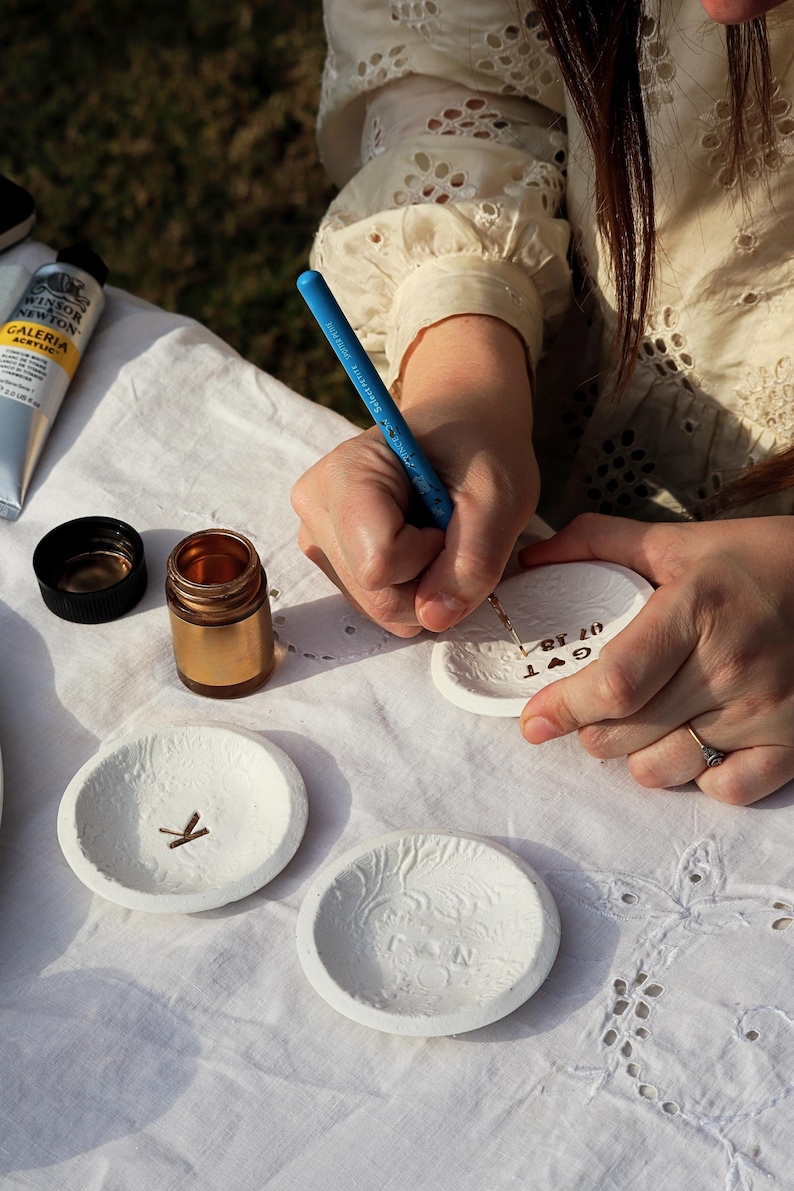 Puede incluir: Una persona est&aacute; rotulando a mano un peque&ntilde;o plato de cer&aacute;mica blanco con un bol&iacute;grafo azul. Otros platos blancos con dise&ntilde;os en relieve y un peque&ntilde;o tarro dorado est&aacute;n sobre un mantel blanco. Tambi&eacute;n es visible un tubo de pintura acr&iacute;lica.