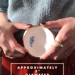 Puede incluir: Un peque&ntilde;o plato de cer&aacute;mica blanca con borde dorado y dise&ntilde;o floral azul. El plato tiene un di&aacute;metro aproximado de 7,6 cm.
