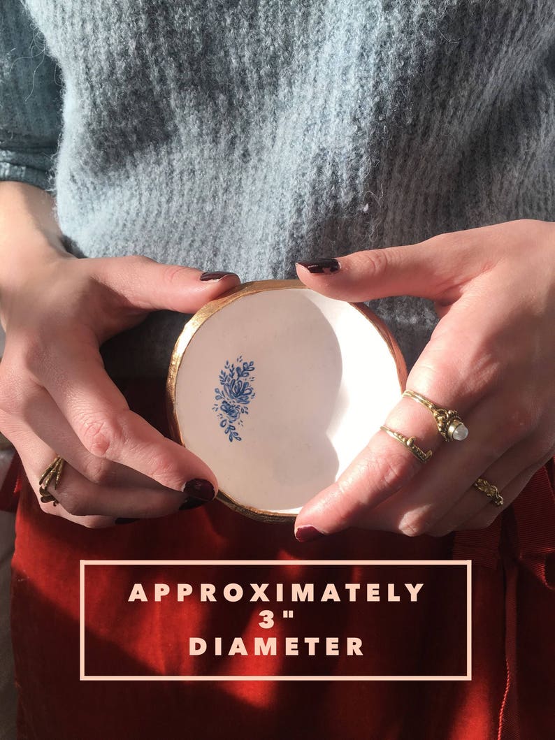 Puede incluir: Un peque&ntilde;o plato de cer&aacute;mica blanca con borde dorado y dise&ntilde;o floral azul. El plato tiene un di&aacute;metro aproximado de 7,6 cm.