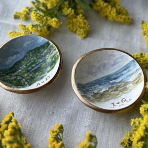 May include: Two small, round, white ceramic dishes with gold rims. One dish has a painted landscape of a mountain scene with the text "A-22" on the bottom. The other dish has a painted landscape of a beach scene with the text "I ♥ G" on the bottom.