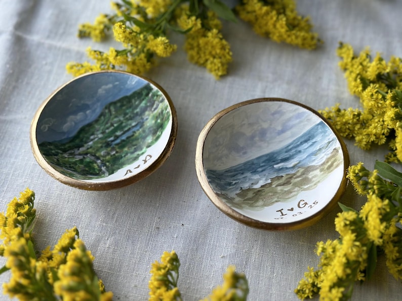 May include: Two small, hand-painted ceramic bowls with gold rims. One bowl depicts a mountain landscape, the other a beach scene. The bowls have personalized initials and dates. Yellow flowers surround the bowls.