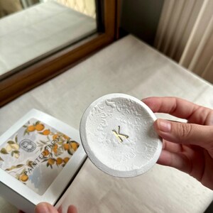 Puede incluir: Un plato peque&ntilde;o, redondo y blanco con un estampado floral y una letra "K" dorada en el centro. El plato est&aacute; sostenido en la mano de una persona. Una caja con un dise&ntilde;o floral est&aacute; en el fondo.