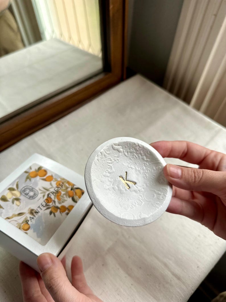 Puede incluir: Un plato peque&ntilde;o, redondo y blanco con un estampado floral y una letra "K" dorada en el centro. El plato est&aacute; sostenido en la mano de una persona. Una caja con un dise&ntilde;o floral est&aacute; en el fondo.