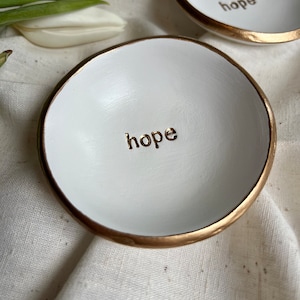 May include: Two white ceramic ring dishes with gold rims. The dishes have the word "hope" written in gold lettering on the inside.