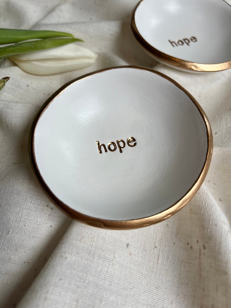 May include: Two white ceramic ring dishes with gold rims. The dishes have the word "hope" written in gold lettering on the inside.