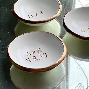 May include: Three white ceramic ring dishes with gold rims. The dishes are personalised with names and dates. The first dish says "M &hearts; J", the second dish says "A &hearts; K 4.8.19", and the third dish is partially visible and says "C &hearts;".