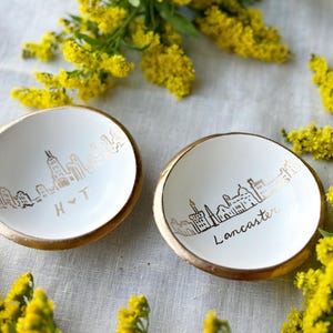May include: Two small, white ceramic dishes with gold rims. Each dish features a gold skyline design and text. One says "H & T", the other says "Lancaster". Yellow flowers surround the dishes.