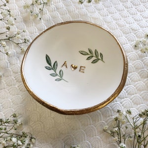 Puede incluir: Plato de cer&aacute;mica blanca para anillos con borde dorado y un coraz&oacute;n dorado y las letras "A E" pintadas en &eacute;l. Hojas verdes est&aacute;n pintadas alrededor de las letras. El plato est&aacute; sobre un fondo blanco.