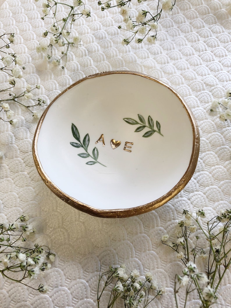 Puede incluir: Plato de cer&aacute;mica blanca para anillos con borde dorado y un coraz&oacute;n dorado y las letras "A E" pintadas en &eacute;l. Hojas verdes est&aacute;n pintadas alrededor de las letras. El plato est&aacute; sobre un fondo blanco.