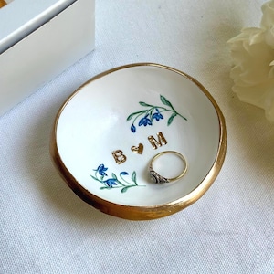 May include: A round white ceramic trinket dish with a metallic gold rim. The interior features hand-painted blue floral sprigs and the gold-colored letters B and M separated by a small heart. A thin gold ring rests inside the dish.