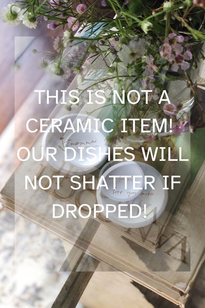 K&ouml;nnte beinhalten: Zwei wei&szlig;e Keramik-Schalen mit handgeschriebenen Botschaften, "Momma" und "Love you more", auf einem Stapel B&uuml;cher. Der Text "This is not a ceramic item! Our dishes will not shatter if dropped!" ist auf dem Bild zu sehen.