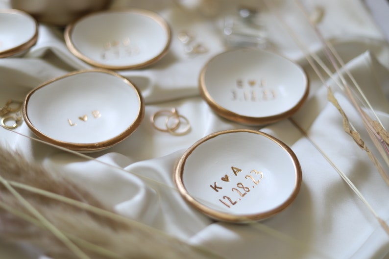 May include: Four white ceramic ring dishes with gold trim. Each dish has a different inscription in gold lettering. The inscriptions are "LOVE", "BOY & GIRL", "K & A 12.28.23", and "12.28.23".